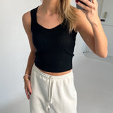 En person iført sort top og hvide joggingbukser tager et spejlbillede i et minimalistisk rum. Personen holder en telefon, og der er ingen tekst i billedet.