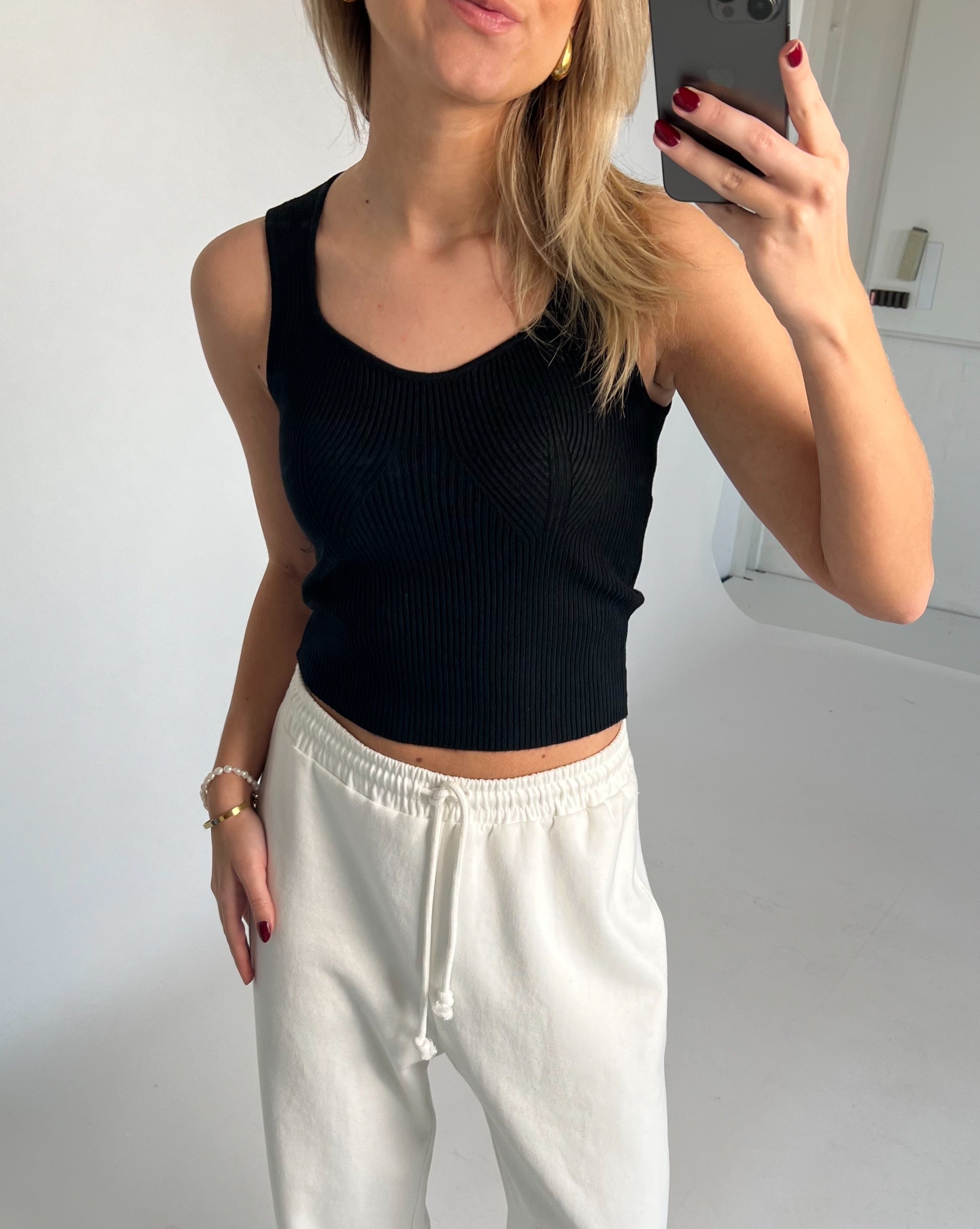 En person iført sort top og hvide joggingbukser tager et spejlbillede i et minimalistisk rum. Personen holder en telefon, og der er ingen tekst i billedet.