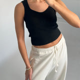 Person iført sort tanktop og hvide joggingbukser tager et spejlbillede, i et rum med lyse vægge. Personen bærer et armbånd af perler på venstre håndled.