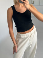 Person iført sort tanktop og hvide joggingbukser tager et spejlbillede, i et rum med lyse vægge. Personen bærer et armbånd af perler på venstre håndled.