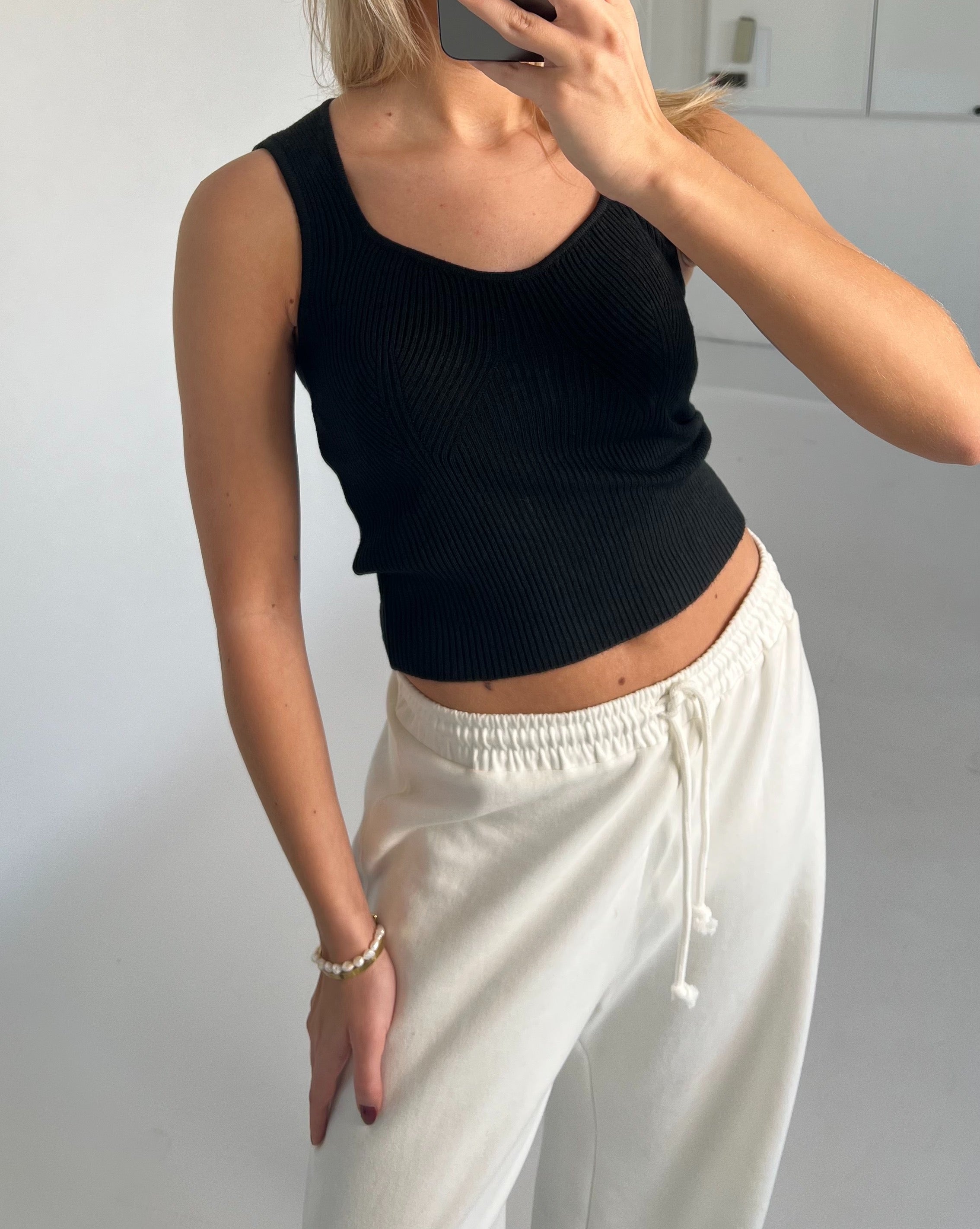 Person iført sort tanktop og hvide joggingbukser tager et spejlbillede, i et rum med lyse vægge. Personen bærer et armbånd af perler på venstre håndled.