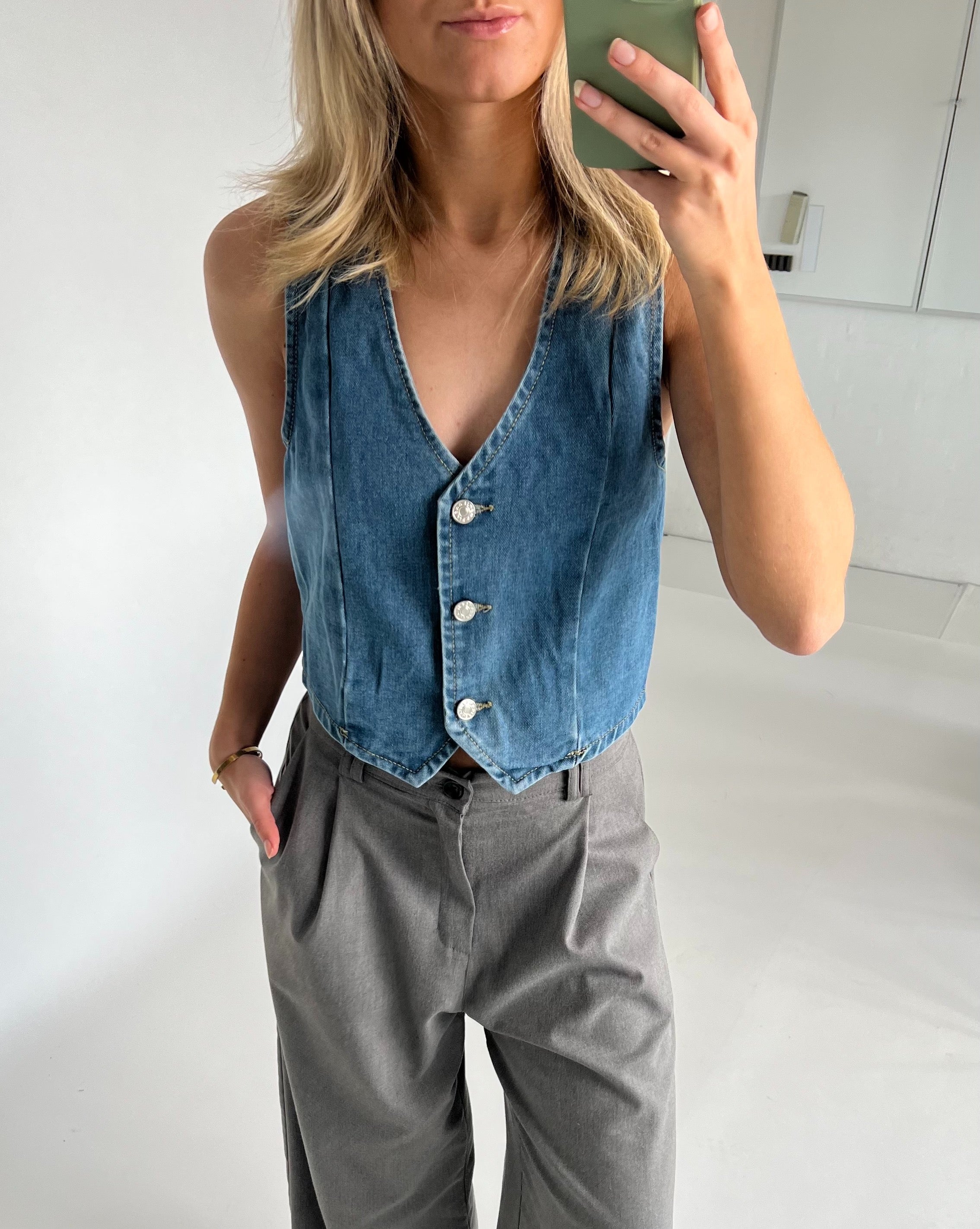 Denimvest bæres af en person, der tager et spejlbillede, handske i grå bukser. Personen står i et lyst rum med hvide vægge.