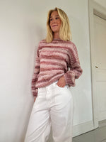 Kvinde iført en stribet, lyserød sweater og hvide bukser står afslappet med hænderne i lommerne mod en hvid væg i et minimalistisk rum.