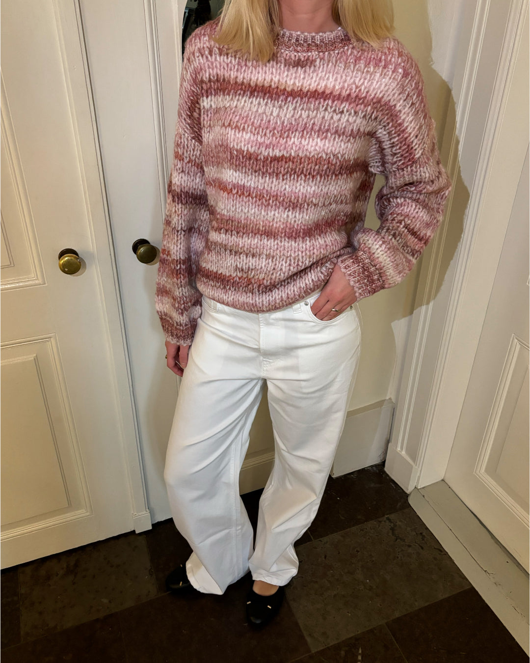 En person bærer en pink-stribet sweater og hvide bukser, står afslappet med højre hånd i lommen i et rum med hvidmalede døre og flisegulv.
