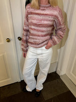 En person bærer en pink-stribet sweater og hvide bukser, står afslappet med højre hånd i lommen i et rum med hvidmalede døre og flisegulv.