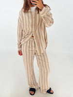 En person iført en beige-stribet pyjamas med afslappet pasform står og tager et spejlbillede med en smartphone, i et lysbelyst rum med hvid baggrund.