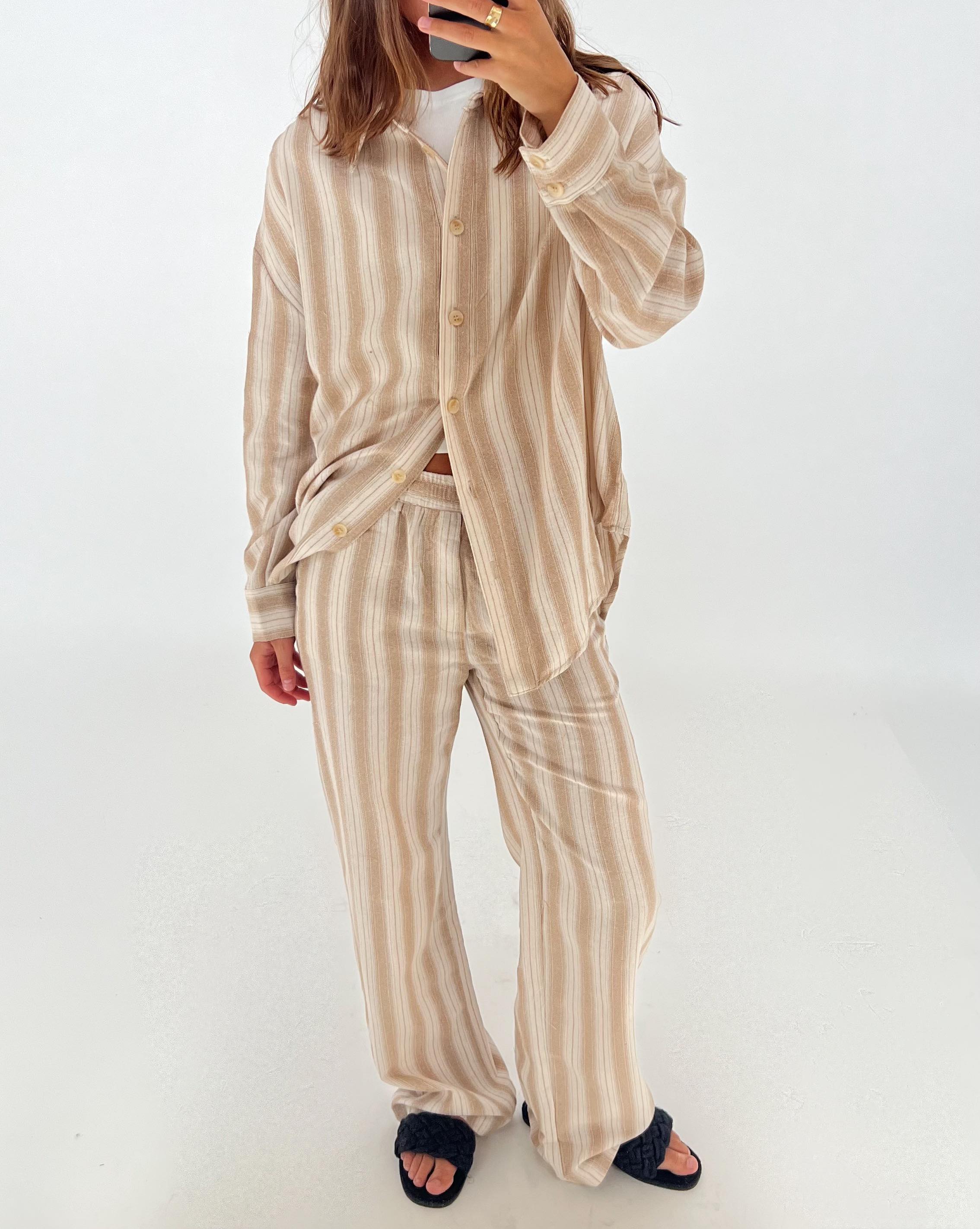 En person iført en beige-stribet pyjamas med afslappet pasform står og tager et spejlbillede med en smartphone, i et lysbelyst rum med hvid baggrund.