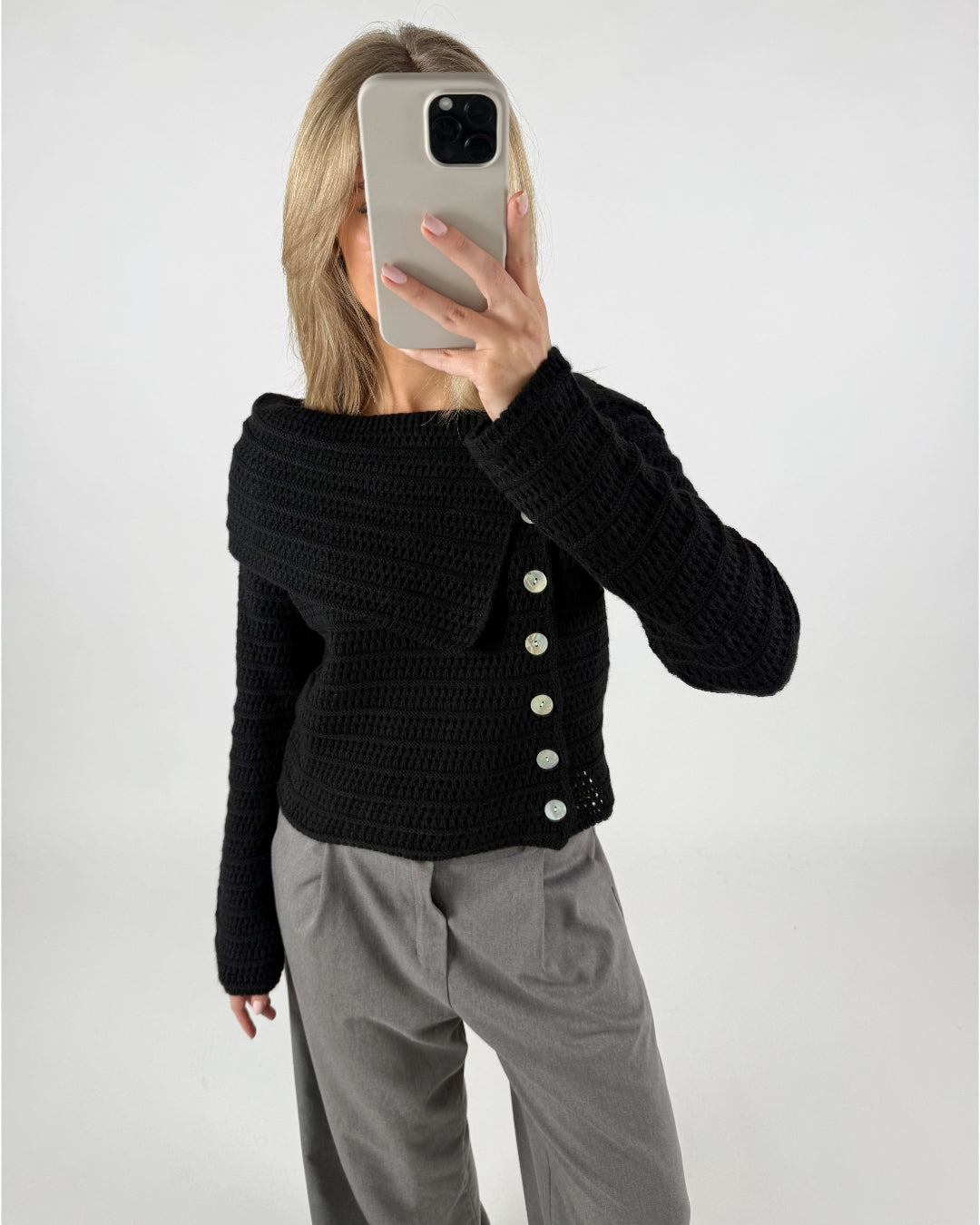 En person holder en mobiltelefon op foran ansigtet i en selfie-position. De bærer en sort strikket trøje med knapper og grå bukser i et lyst rum.