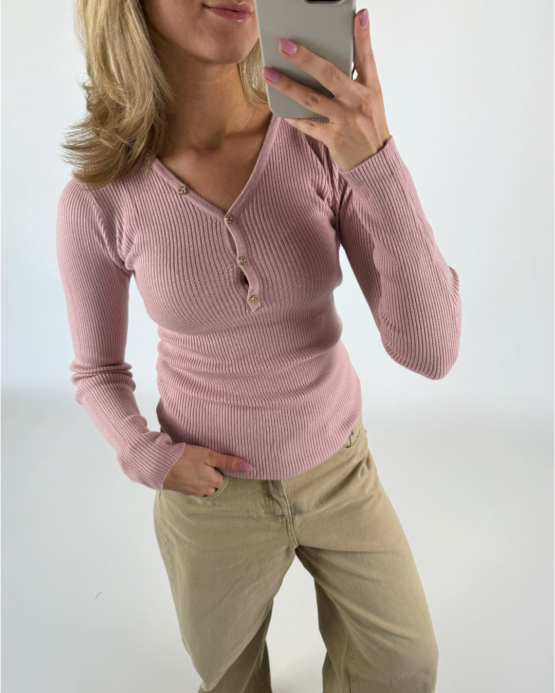Person tager en selfie, iført en lyserød ribstriktrøje og beige bukser. Hånden er i bukselommen, mens telefonen holdes foran et lyst baggrundsmiljø.