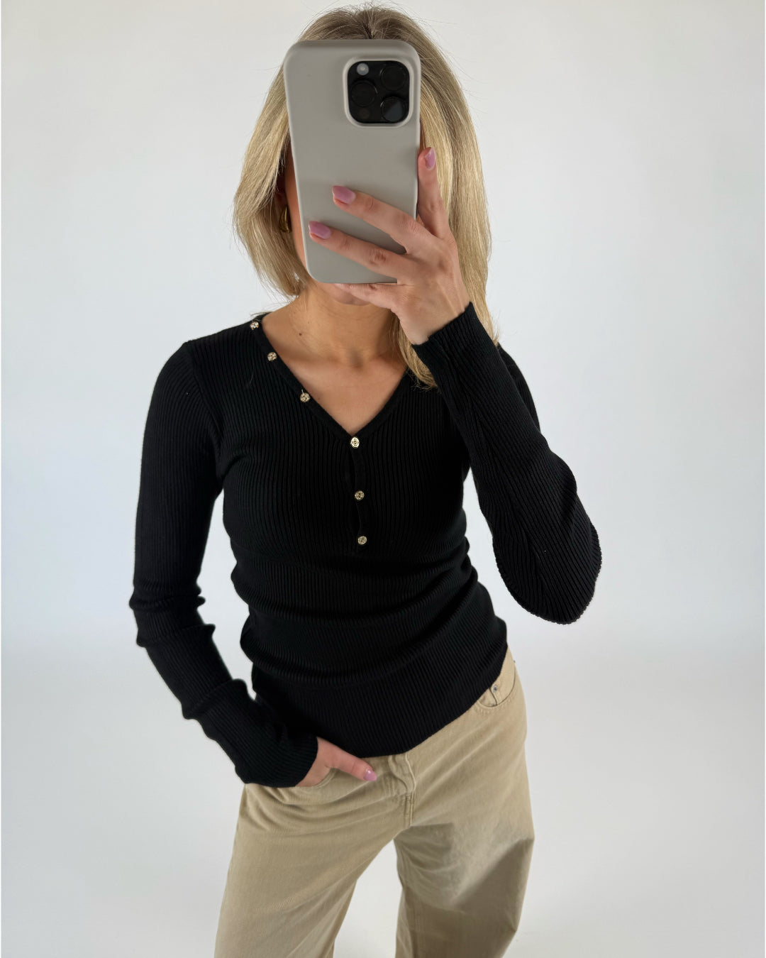 Person tager et spejlbillede med en smartphone og skjuler ansigtet. Personen bærer en sort trøje med knapper og beige bukser i et neutralt oplyst rum.