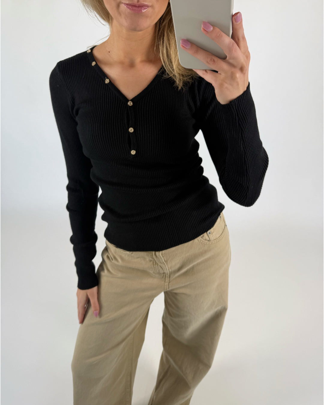En person iført en sort trøje med knapper tager en selfie i et spejl. Vedkommende har beige bukser på, med en neutral baggrund.