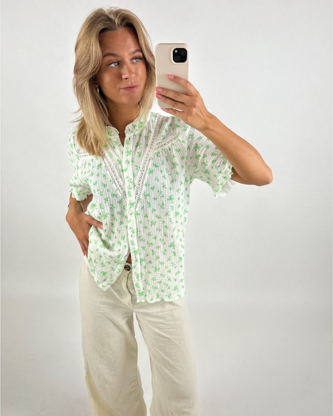 Kvinde iført en hvid bluse med grønt blomstermønster tager en selfie. Hun står afslappet i enkle, lyse bukser mod en hvid baggrund.