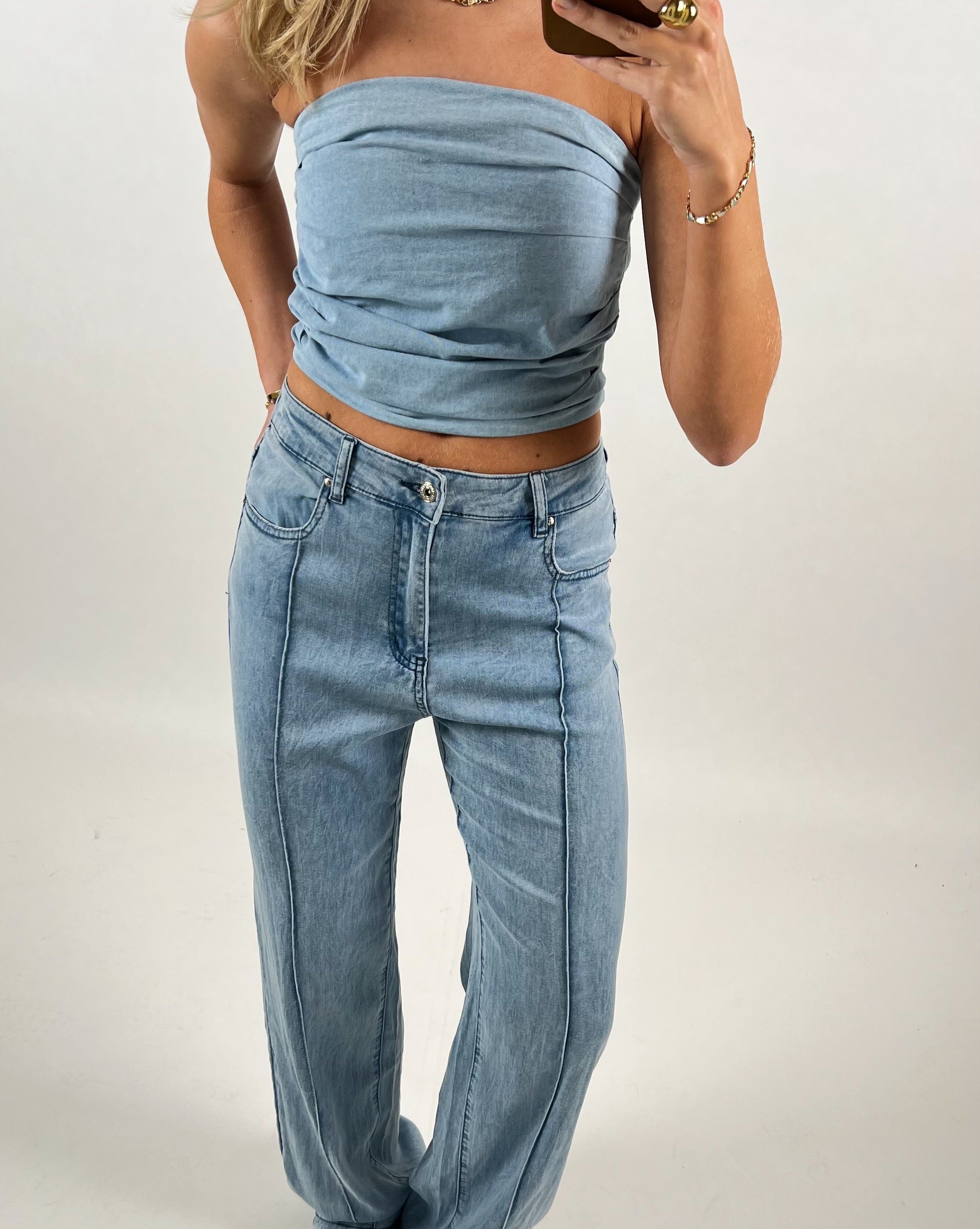 En person iført en stropløs top og jeans poserer, mens de tager et spejlbillede med en smartphone. Baggrunden er enkle og hvid, hvilket fremhæver tøjet.