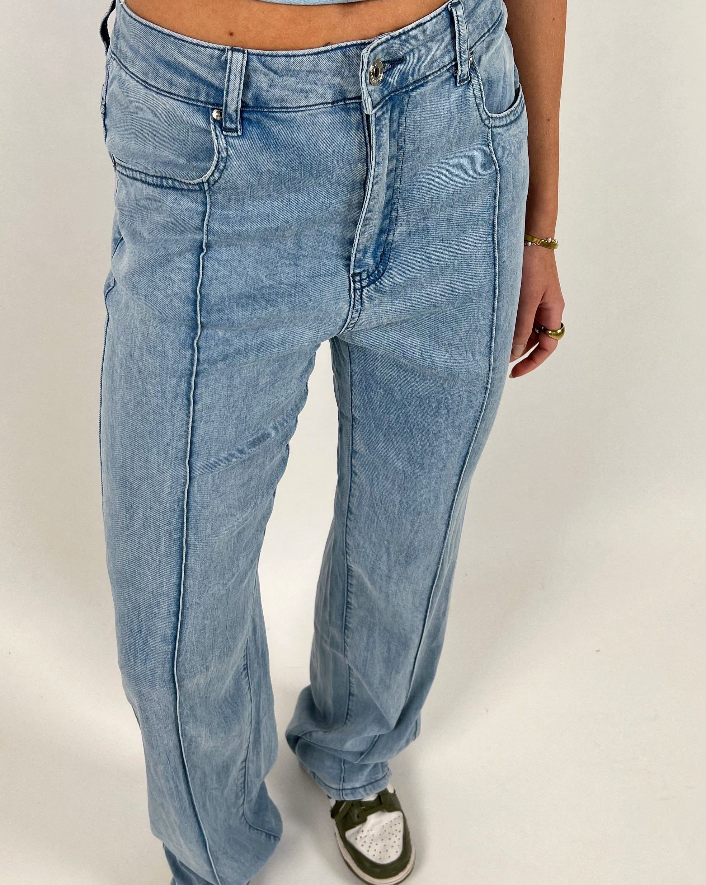 Lyseblå jeans bæres af en person, der står med armen let sænket. Personen har en lysebrun hudtone, iført en grøn sneaker med hvide detaljer. Gulvet er hvidt.