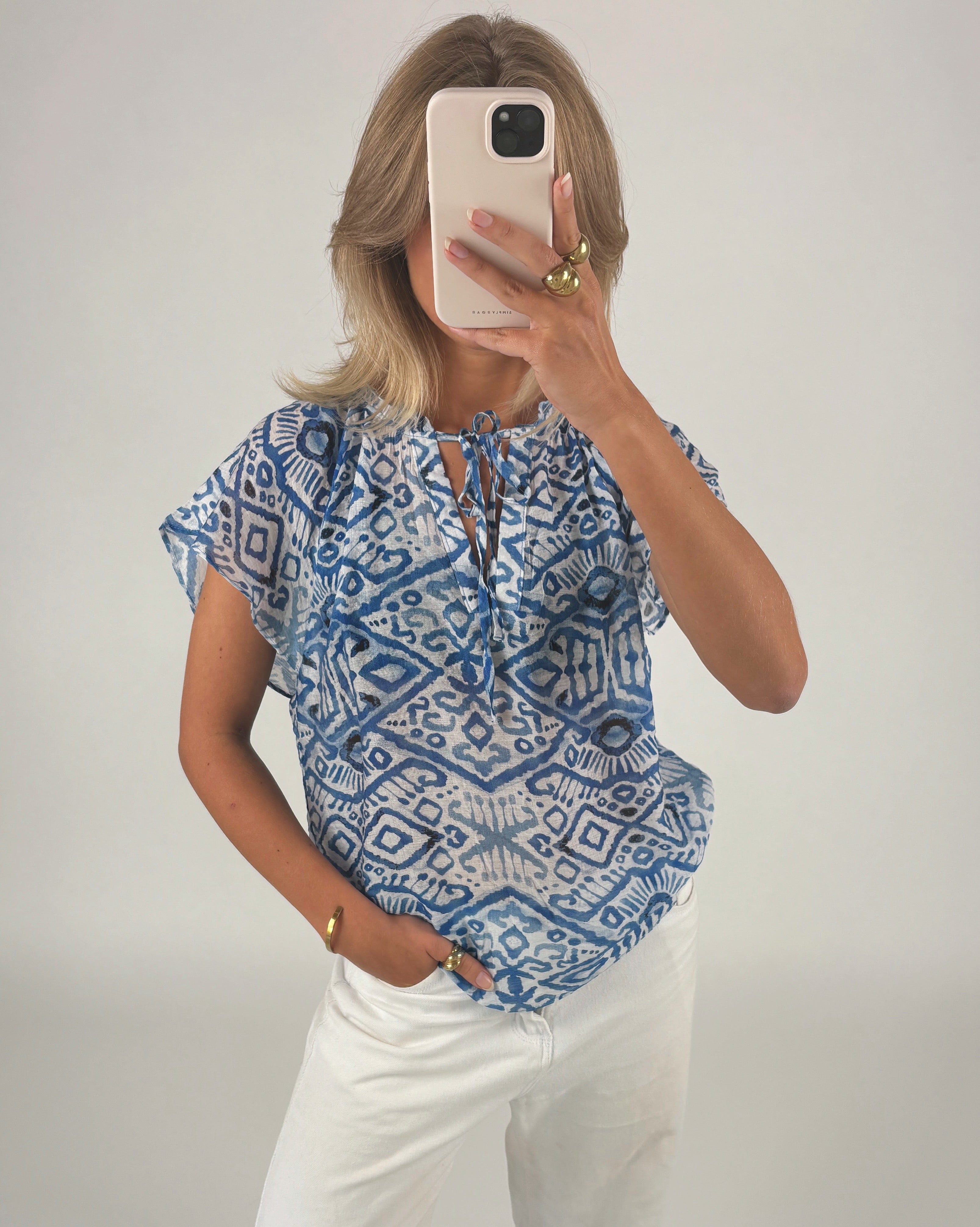 Person tager en selfie med mobiltelefon, iført en blå-hvid mønstret bluse og hvide bukser i et neutralt rum. Hånden har flere smykker, herunder ringe og et armbånd.