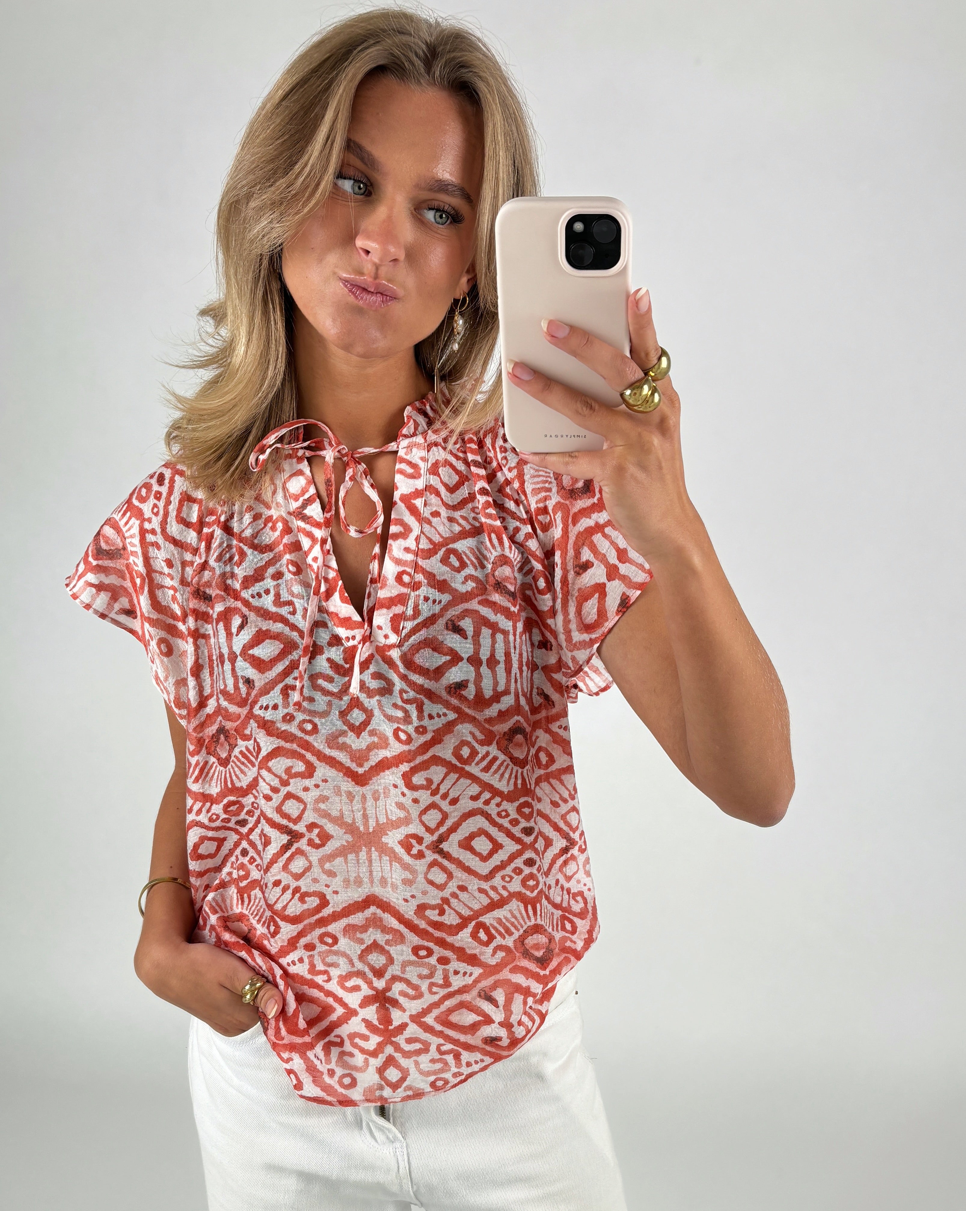 Kvinde tager selfie iført mønstret rød og hvid bluse og hvide bukser. Hun holder en beige telefon i hånden med ringe, i en lys, neutral baggrund.