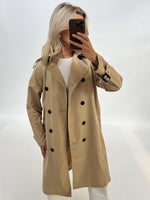 En person iført en beige trenchcoat tager en selfie med en smartphone. Personen står i et lys oplyst rum med en hvid baggrund, iført en hvid trøje under frakken.