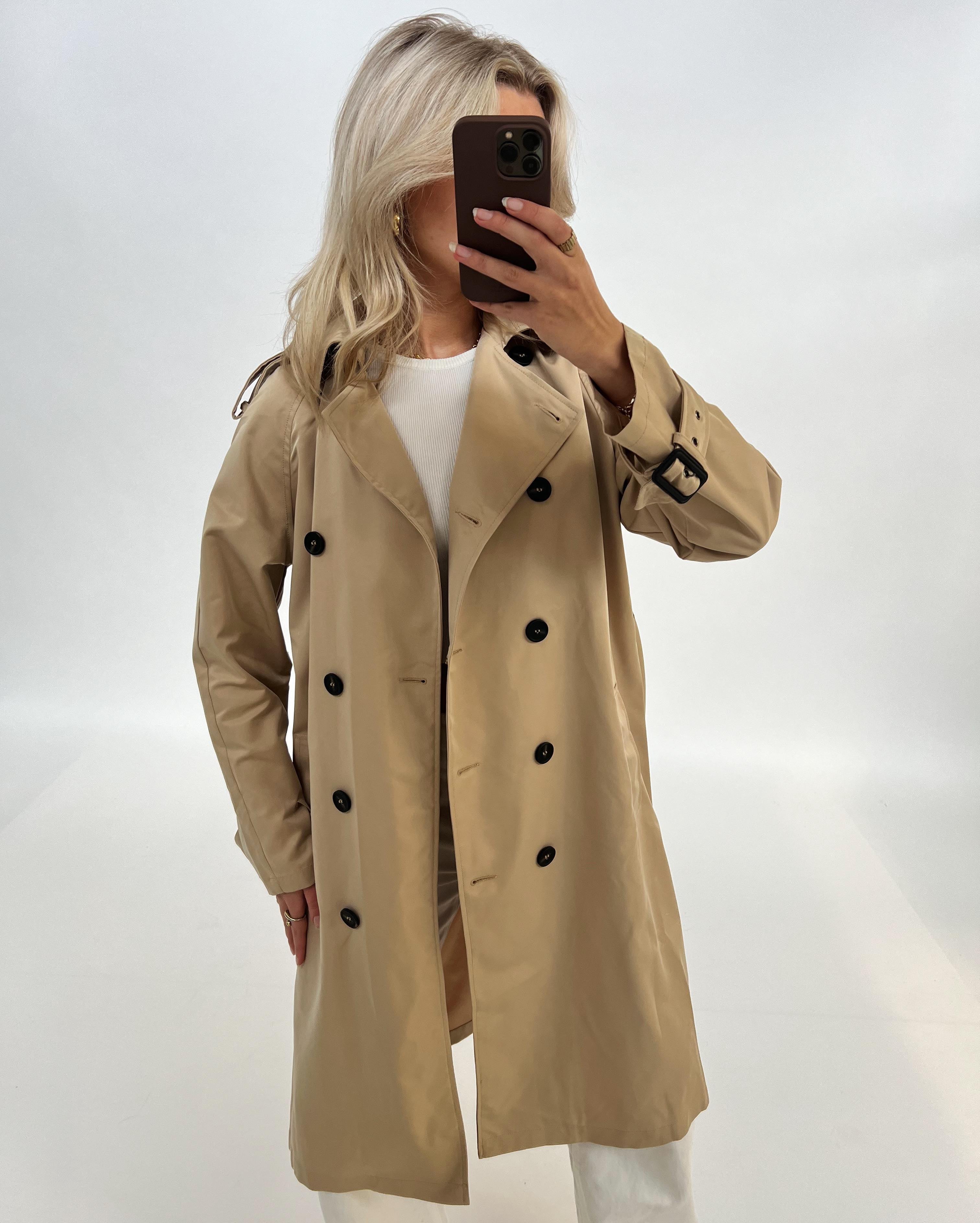 En person iført en beige trenchcoat tager en selfie med en smartphone. Personen står i et lys oplyst rum med en hvid baggrund, iført en hvid trøje under frakken.