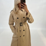 En person iført en beige trenchcoat tager et spejlbillede med en smartphone. Personen står i et lyst, neutralt rum uden synlige mønstre eller genstande.