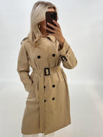 En person iført en beige trenchcoat tager et spejlbillede med en smartphone. Personen står i et lyst, neutralt rum uden synlige mønstre eller genstande.