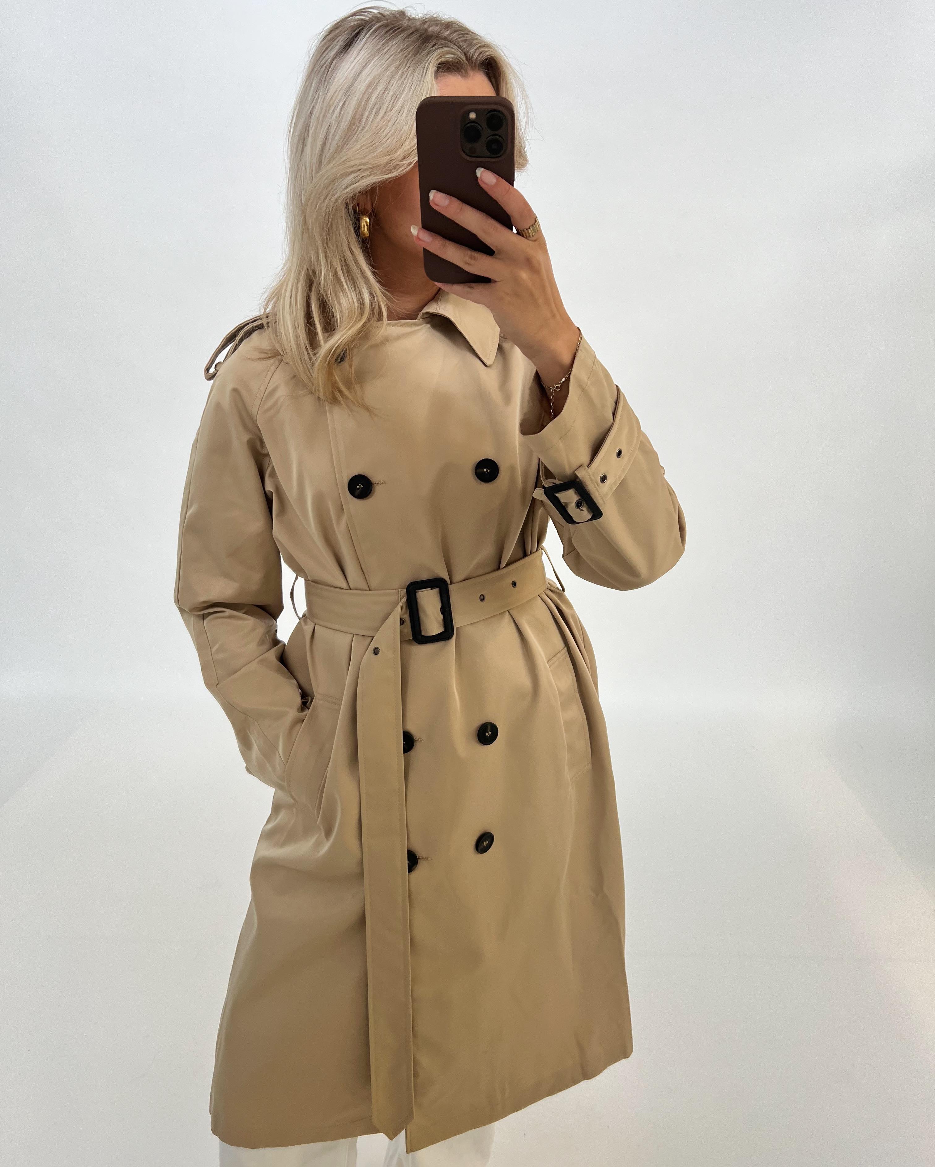 En person iført en beige trenchcoat tager et spejlbillede med en smartphone. Personen står i et lyst, neutralt rum uden synlige mønstre eller genstande.
