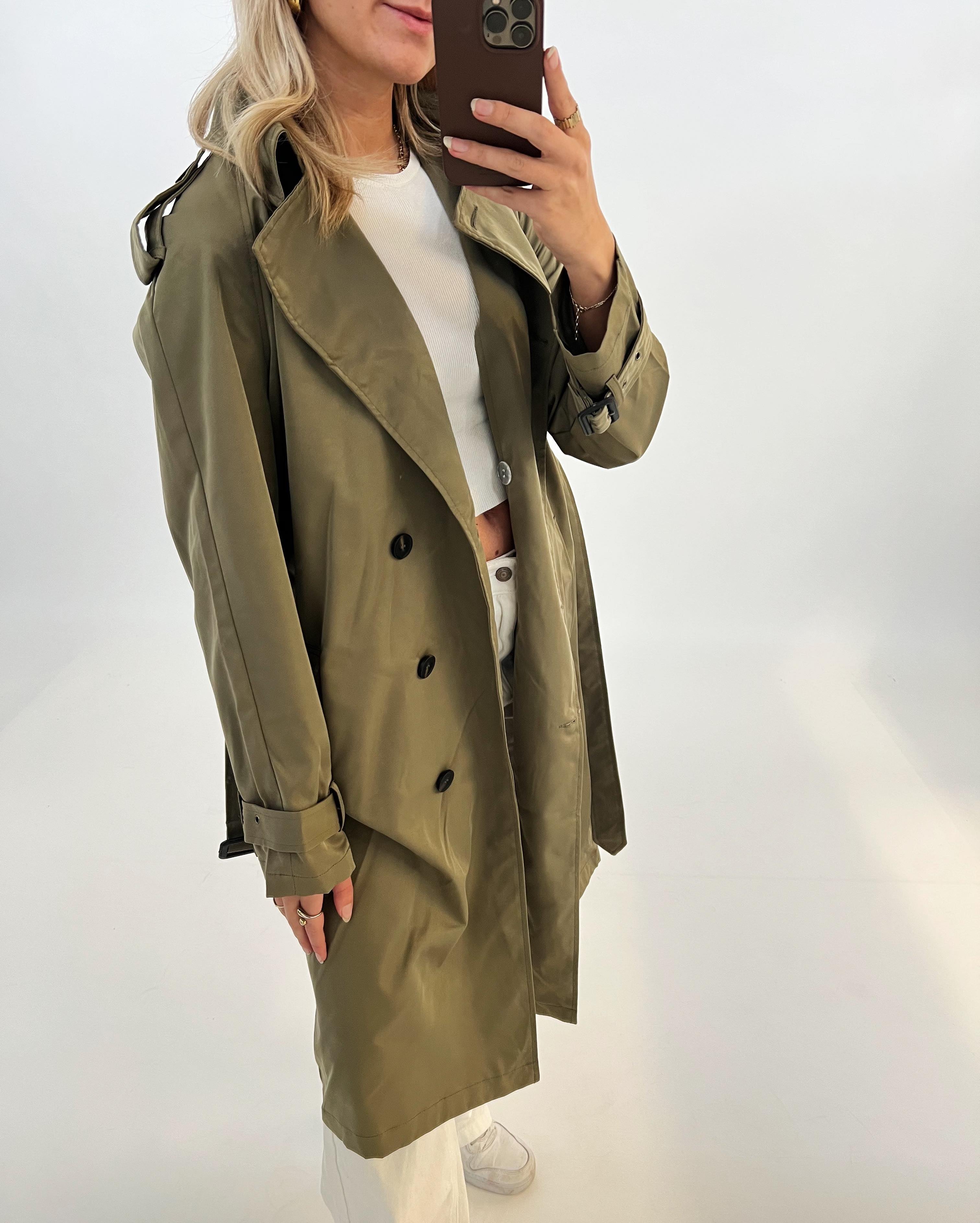 En person iført en grøn trenchcoat tager en selfie i et spejl. Personen bærer også en hvid trøje og bukser i en lys, minimalistisk baggrund.