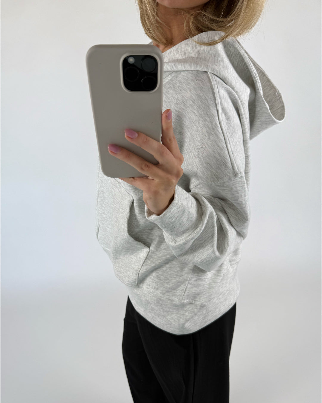 En person iført en grå hættetrøje og sorte bukser står i et lyst rum og tager et spejlbillede med en smartphone.