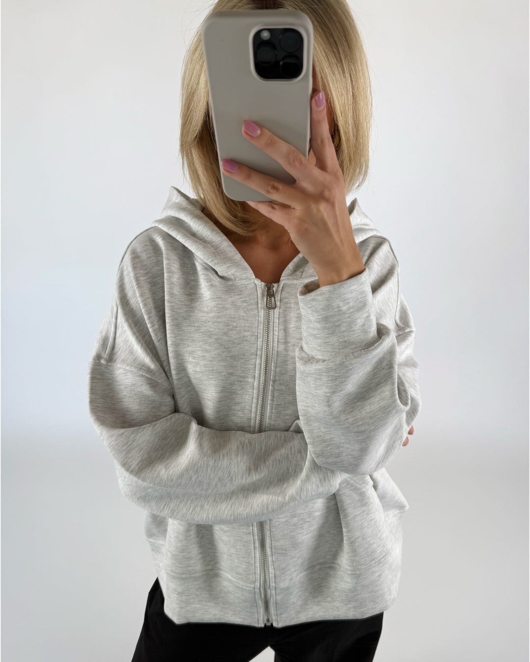 Person i grå hættetrøje holder en smartphone foran ansigtet, hvilket dækker det. Baggrunden er lys og neutral, hvilket fremhæver personen og telefonen.