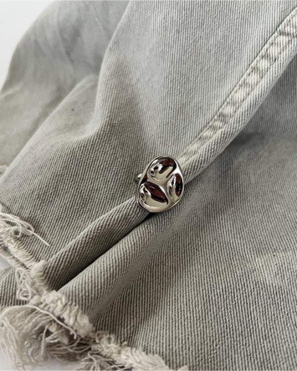 En sølvfarvet broche sidder på grå, let slidt denimstof. Brochen har blanke, blødt formede overflader og lyser op i kontrast til materialets matte tekstur.