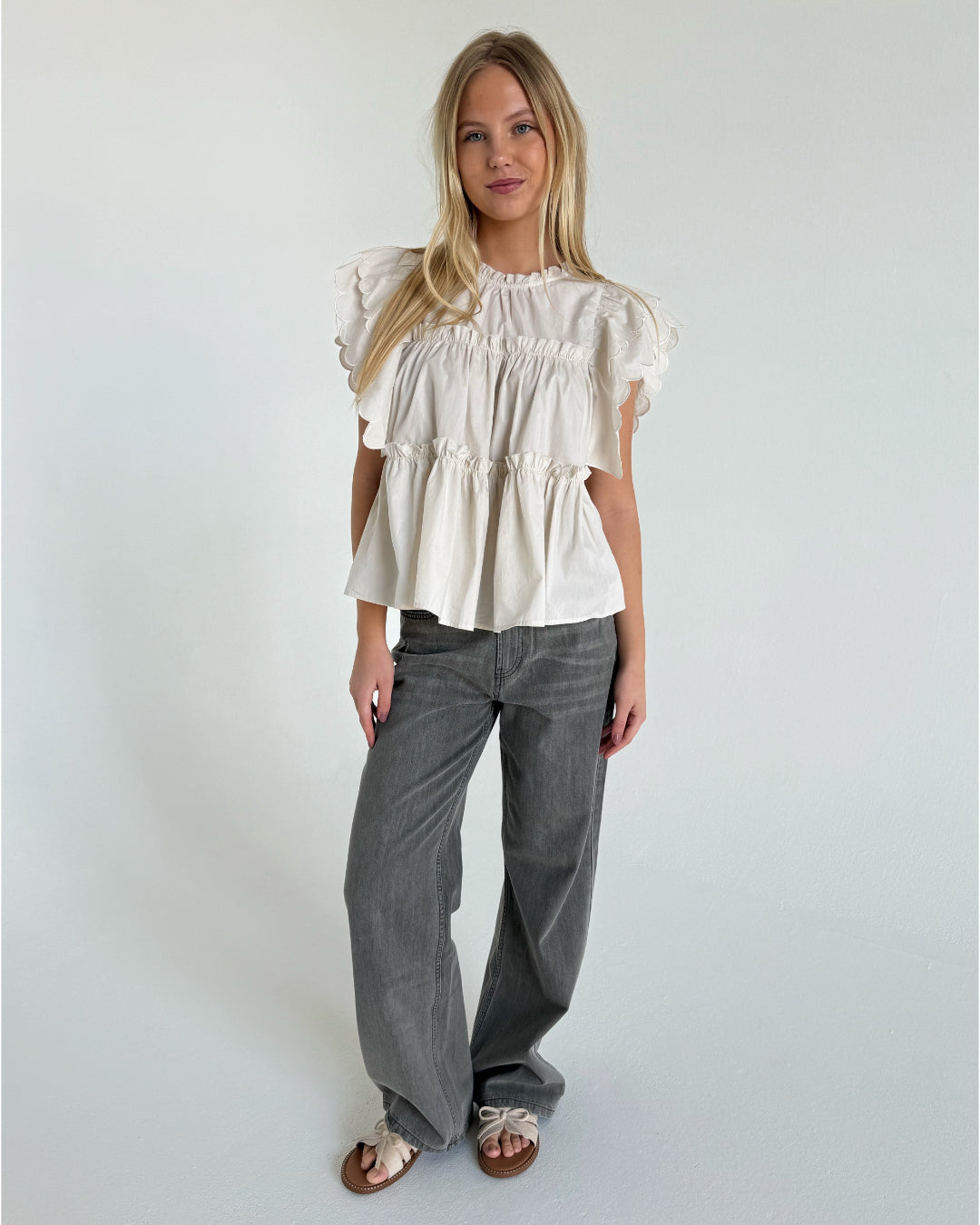 Kvinde iført hvid, rynket, kortærmet bluse og grå, løstsiddende jeans står på hvidt gulv mod en neutral bagvæg, ser afslappet fremad.