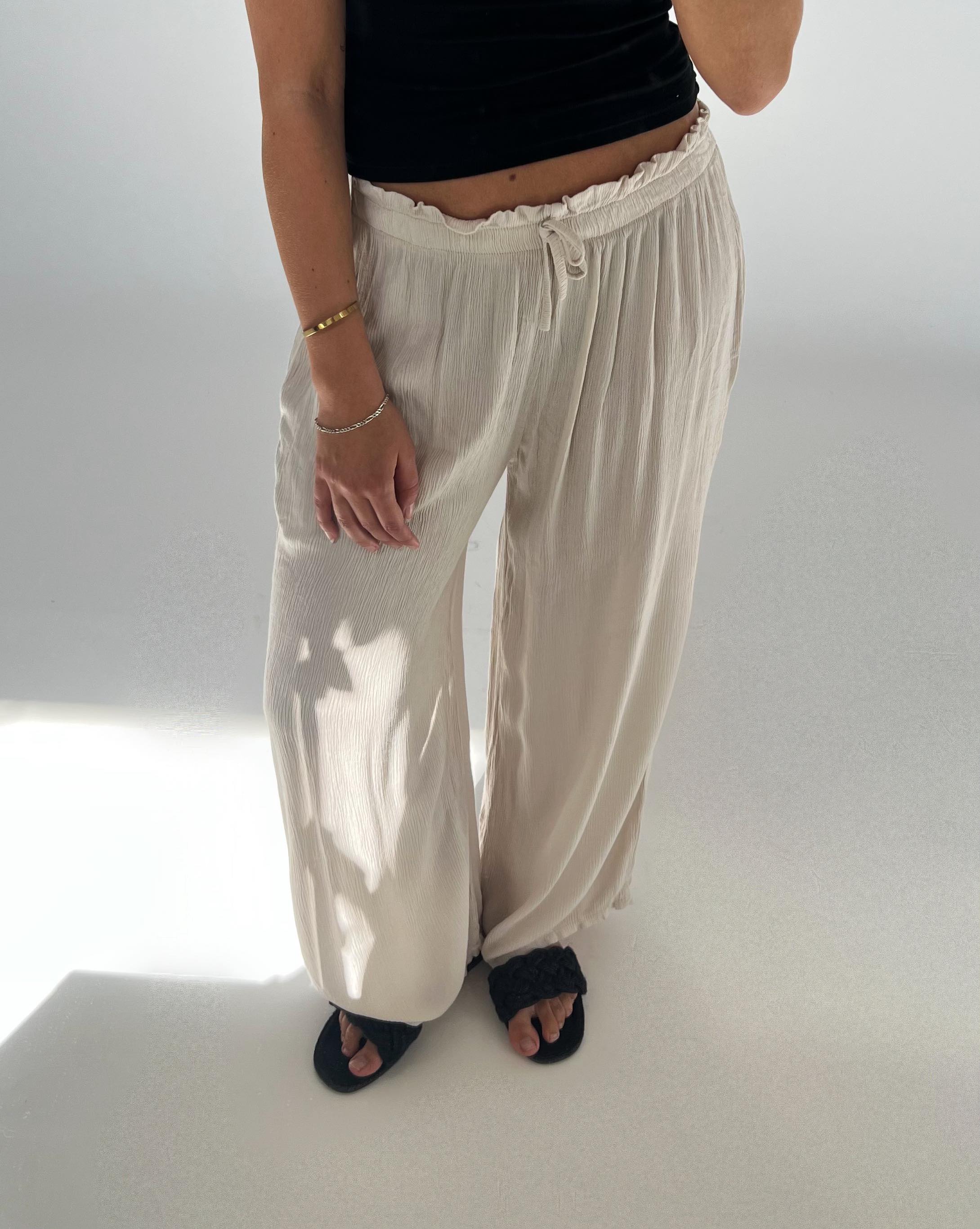 En person iført sorte sandaler og beige, løse bukser står afslappet på et lyst gulv. Personen bærer en sort top og et guldfarvet armbånd.