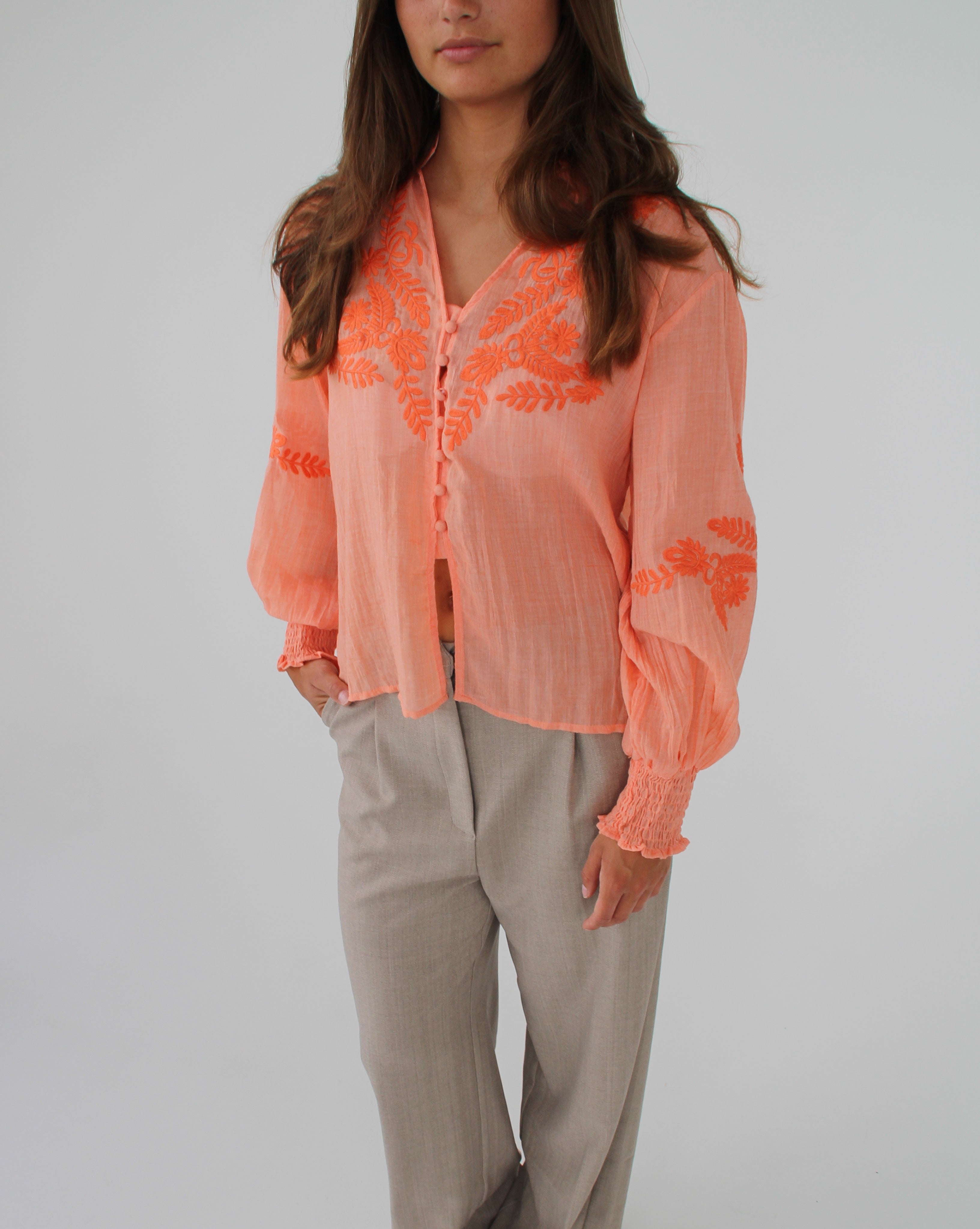En person bærer en lys orange bluse med blomsterbroderi og beige bukser, står mod en hvid baggrund.