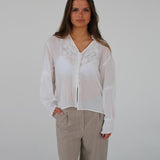 En person står i en hvid bluse med broderede detaljer og beige bukser, med hænderne i lommerne, poserer mod en neutral, lys baggrund.