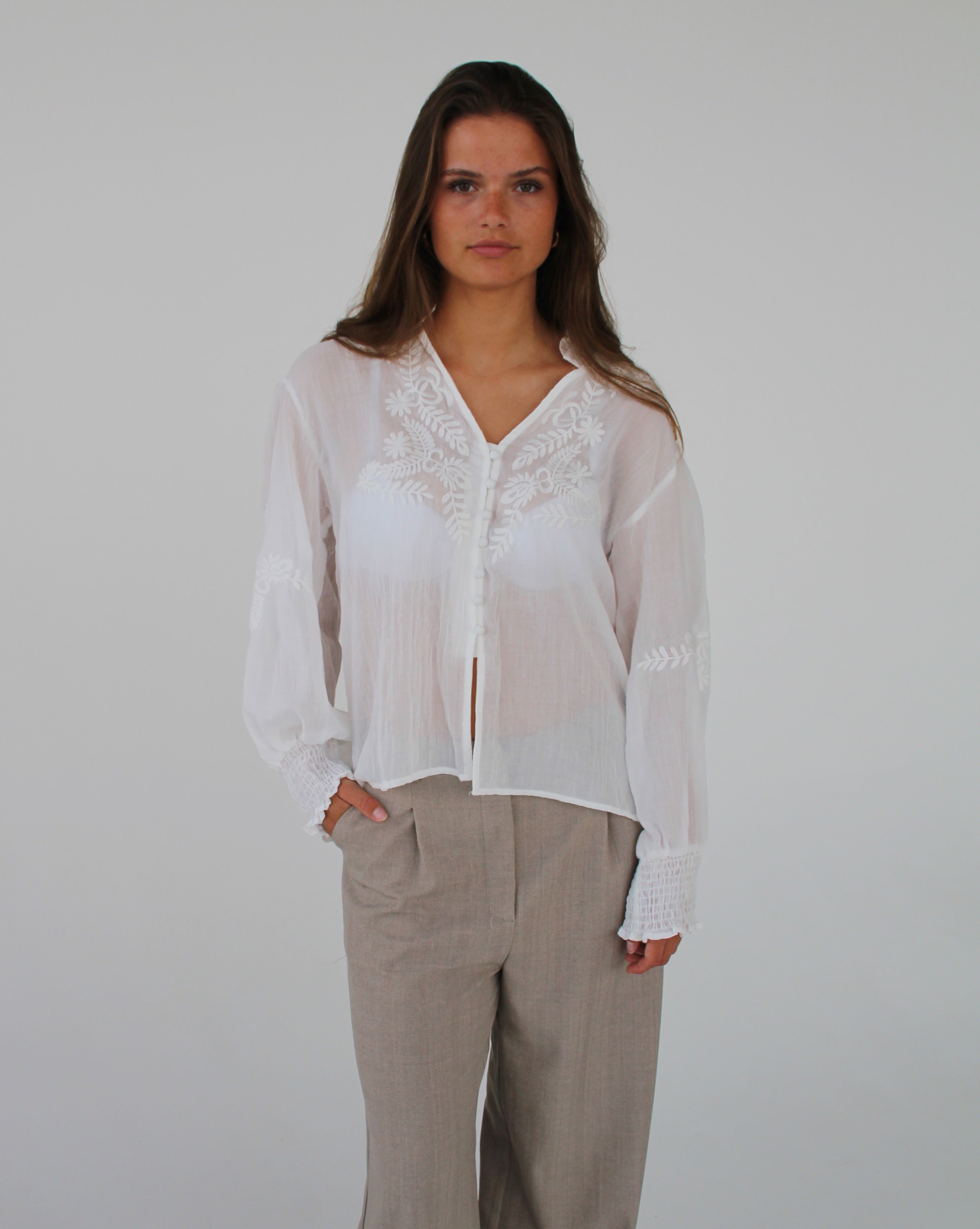 En person står i en hvid bluse med broderede detaljer og beige bukser, med hænderne i lommerne, poserer mod en neutral, lys baggrund.