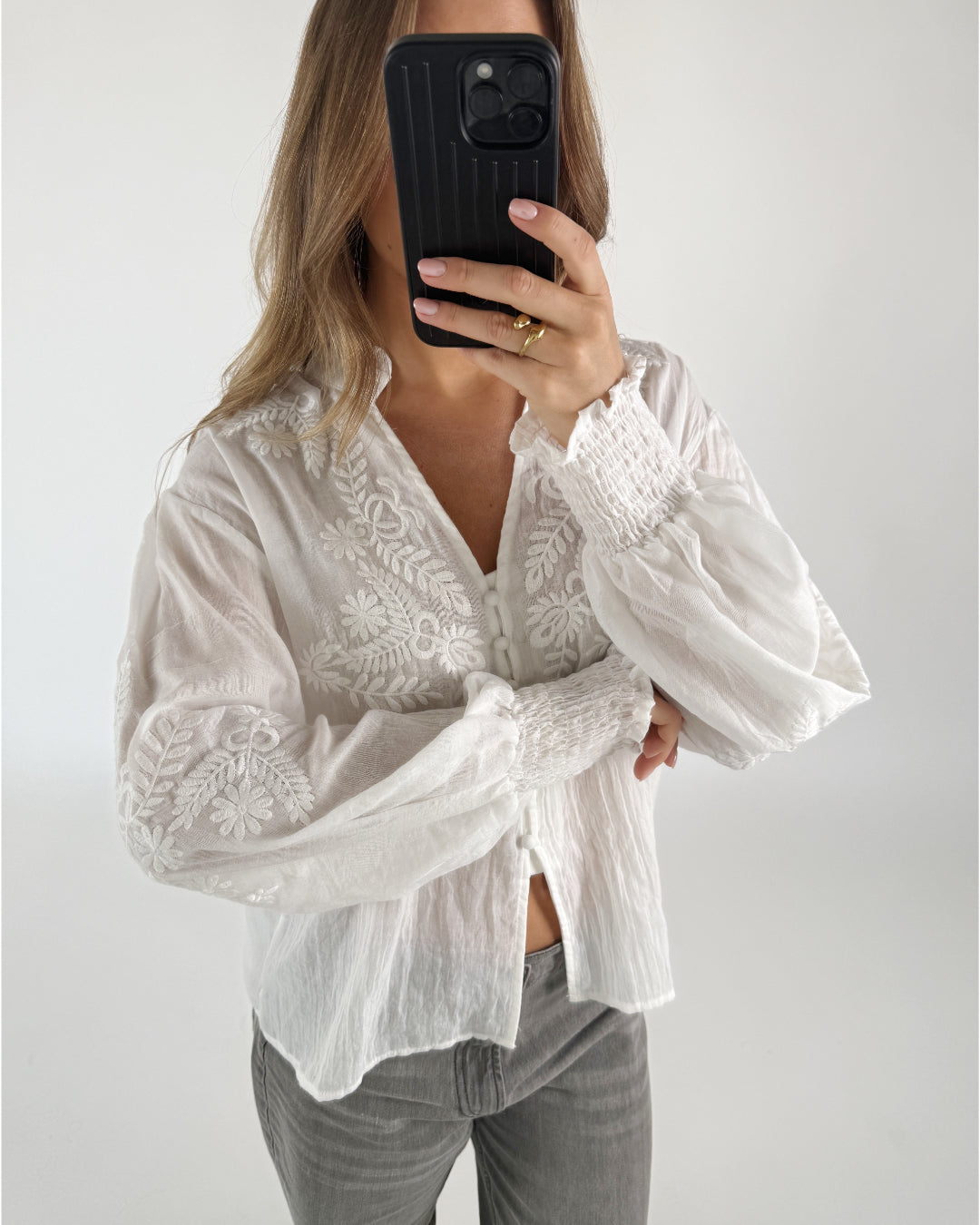 En person iført en hvid, broderet bluse tager et selfie med en sort telefon. Personen står i et lyst rum uden synlige møbler.