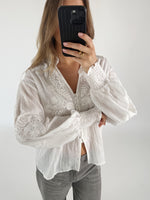 En person iført en hvid, broderet bluse tager et selfie med en sort telefon. Personen står i et lyst rum uden synlige møbler.