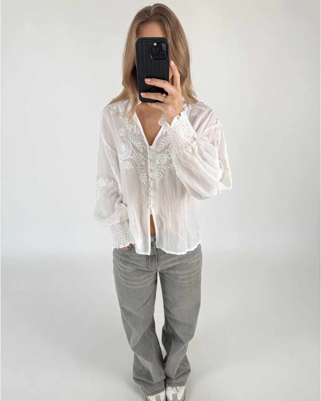 En person iført en hvid, broderet bluse og grå bukser holder en mobiltelefon op foran ansigtet i et neutralt, lyst rum.