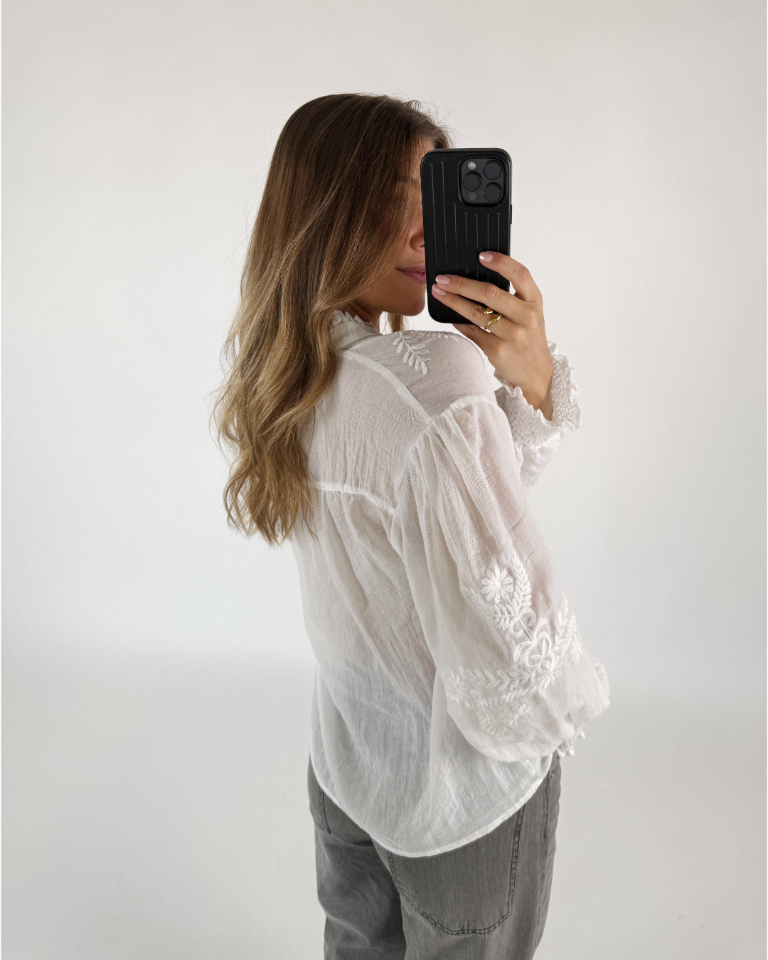 En person tager et selfie med en mobiltelefon, iført en hvid, broderet bluse. Deres lange hår falder over skuldrene, og personen står i et rum med en hvid baggrund.