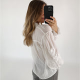En person tager et selfie med en mobiltelefon, iført en hvid, broderet bluse. Deres lange hår falder over skuldrene, og personen står i et rum med en hvid baggrund.
