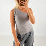 En person iført gråt asymmetrisk bluse og bukser tager en selfie med en smartphone i et minimalistisk rum. Personen har blondt hår og bærer guldsmykker.