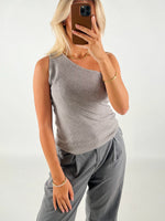 En person iført gråt asymmetrisk bluse og bukser tager en selfie med en smartphone i et minimalistisk rum. Personen har blondt hår og bærer guldsmykker.