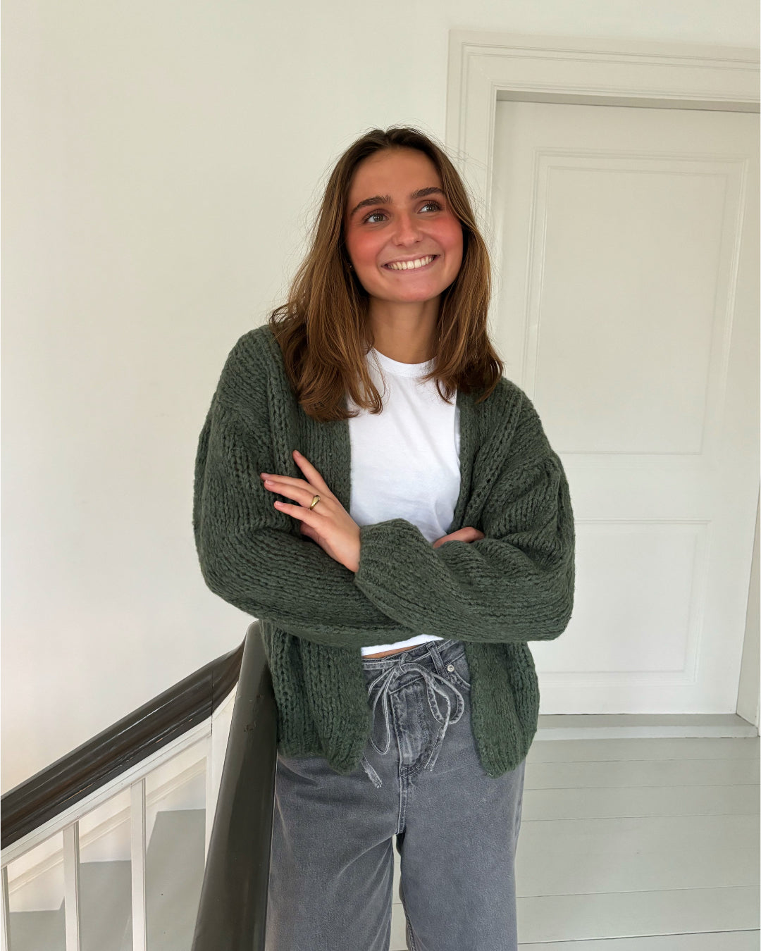 En person iført en hvid T-shirt og grøn strikket cardigan står smilende i en lys gang nær en hvid dør, med armene let krydsede.