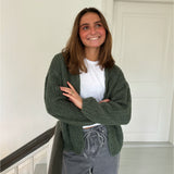 En person iført en hvid T-shirt og grøn strikket cardigan står smilende i en lys gang nær en hvid dør, med armene let krydsede.