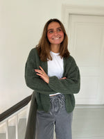 En person iført en hvid T-shirt og grøn strikket cardigan står smilende i en lys gang nær en hvid dør, med armene let krydsede.