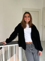 En person smiler og hviler sin arm på et gelænder. Personen bærer en sort trøje over en hvid t-shirt og grå bukser. Rummet har hvide vægge og døre.