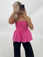 En person tager en selfie iført en pink stropløs top og sorte bukser, mens de står i et lyst rum med en neutral baggrund.