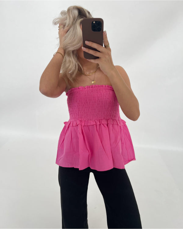 En person tager en selfie iført en pink stropløs top og sorte bukser, mens de står i et lyst rum med en neutral baggrund.