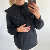 En person i sort skjorte med stor krave tager en selfie med en brun telefon i et lyst rum. Personen har rødmalede negle og guldsmykker.