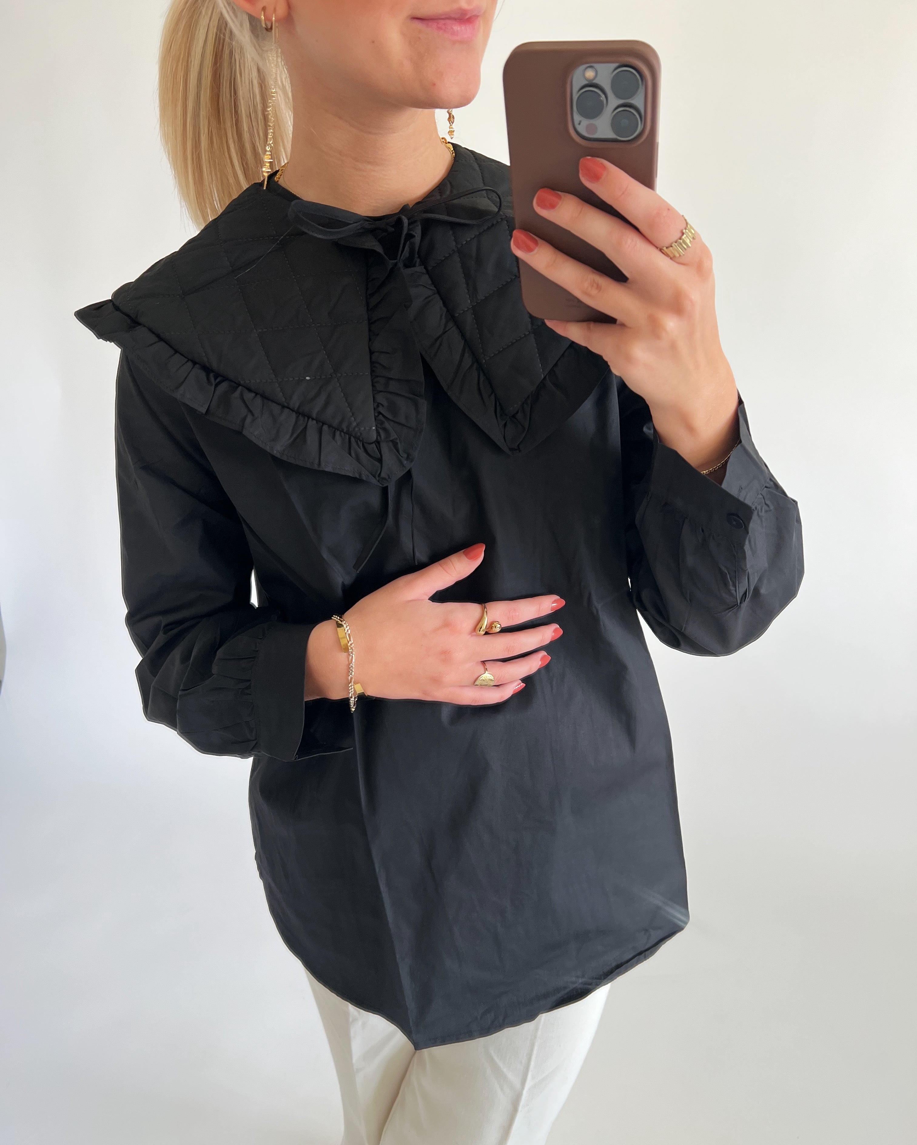 En person i sort skjorte med stor krave tager en selfie med en brun telefon i et lyst rum. Personen har rødmalede negle og guldsmykker.
