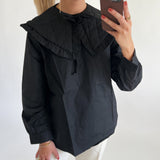 En person, iført sort bluse med stor krave, tager en selfie i spejlet med en mobiltelefon. Personen bærer også øreringe og flere ringe, og har lyst hår.
